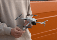 Person in a beige hoodie holds a gray quadcopter drone with four propellers in front of an orange vehicle camera facing forward